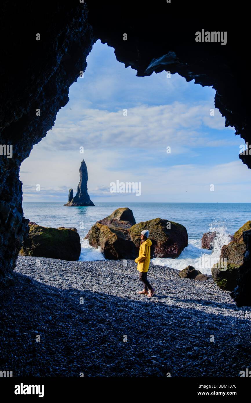 Eine Frau, die in einer felsigen Höhle steht, bewundert den atemberaubenden Strand von Reynisfjara. Majestätisch erheben sich die dramatischen Meeresstapel im Hintergrund, während die Wellen in Island gegen die Küste krachen Stockfoto