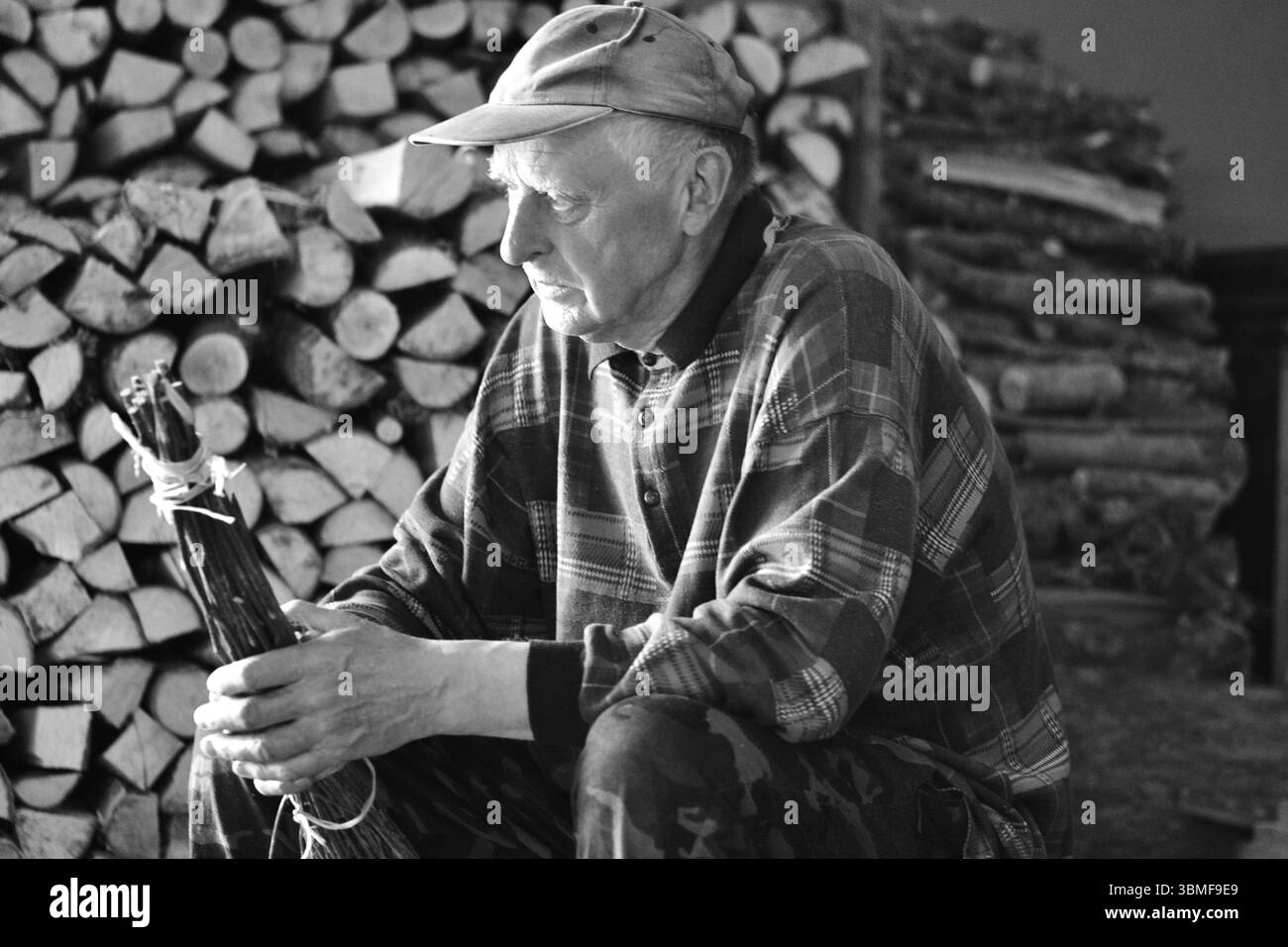 Schwarz-weiß-Foto eines älteren Bauern, der vor gestapeltem Brennholz sitzt und einen handgefertigten Bodenpinsel hält. Rustikale Szene mit traditionellem rura Stockfoto