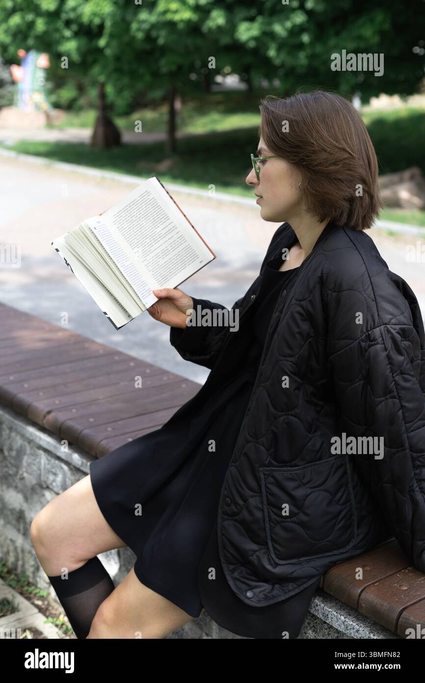 Eine junge Frau sitzt ruhig auf einer Parkbank, in ein Buch eingetaucht, umgeben von hohen grünen Bäumen. Sie trägt einen schwarzen Rock und eine schwarze Jacke, verloren in der Ruhe o Stockfoto