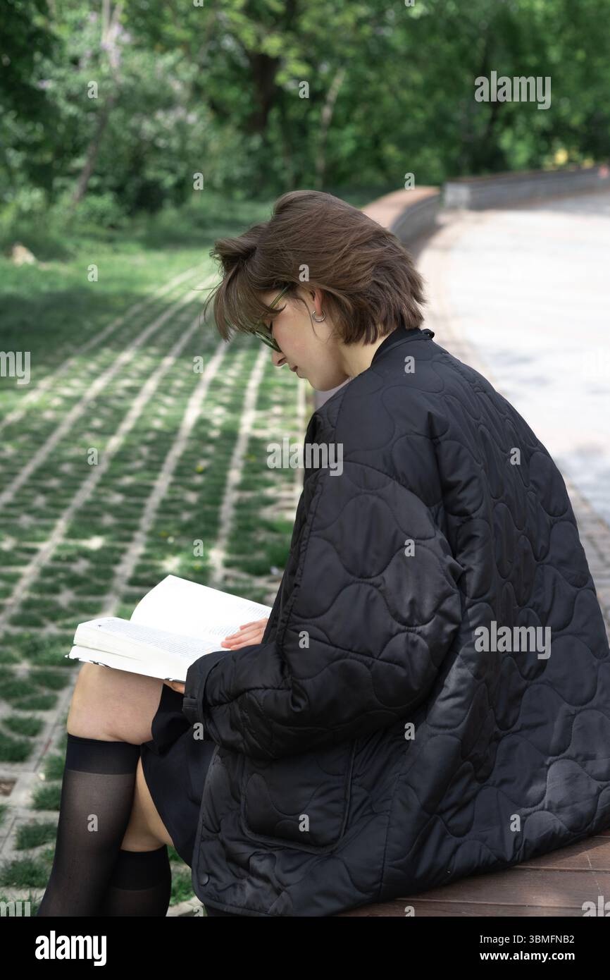 Allein im sanften Schatten der Bäume liest eine Frau in schwarzem Rock und Jacke einen friedlichen Moment auf einer Holzbank im Stadtpark. Stockfoto