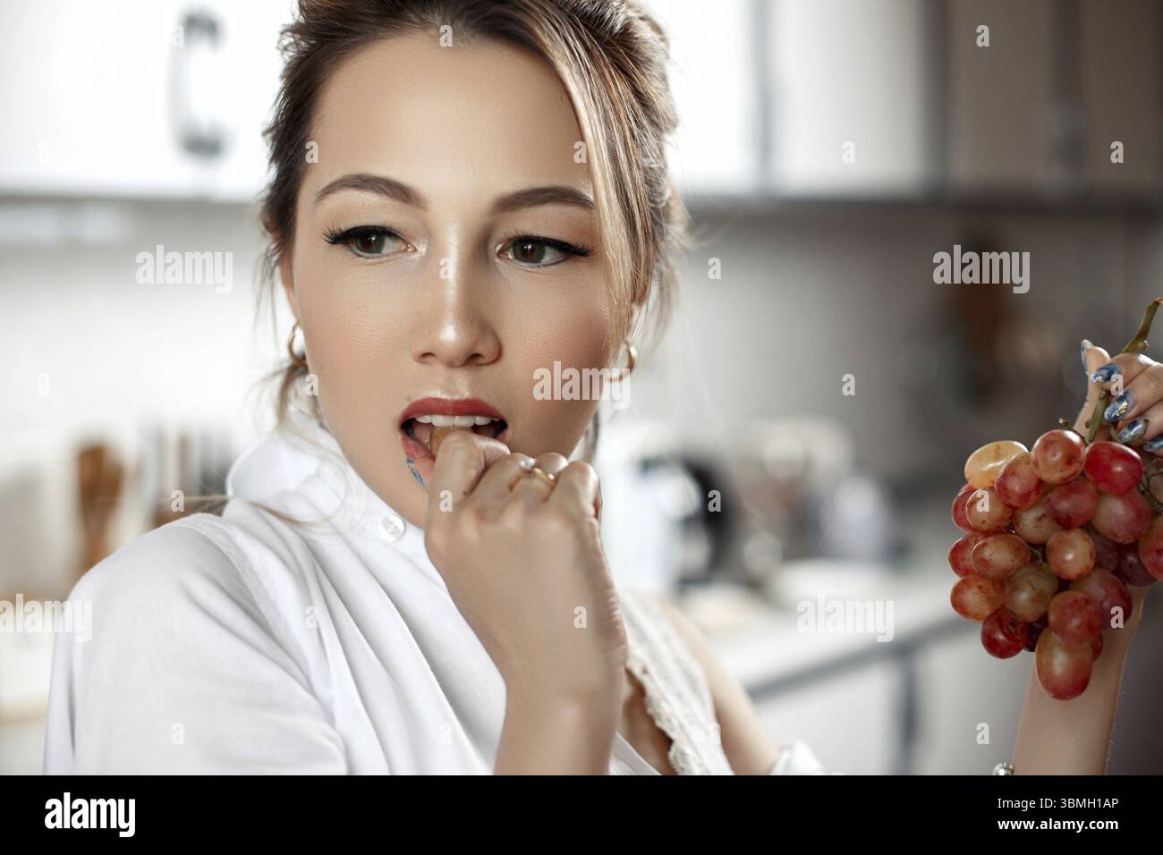 Junge schöne asiatische Frau in beigen Spitzen-Shorts, weißes Hemd, das Trauben in der Küche isst. Selektiver Fokus: Ein kleiner Fokusbereich Stockfoto