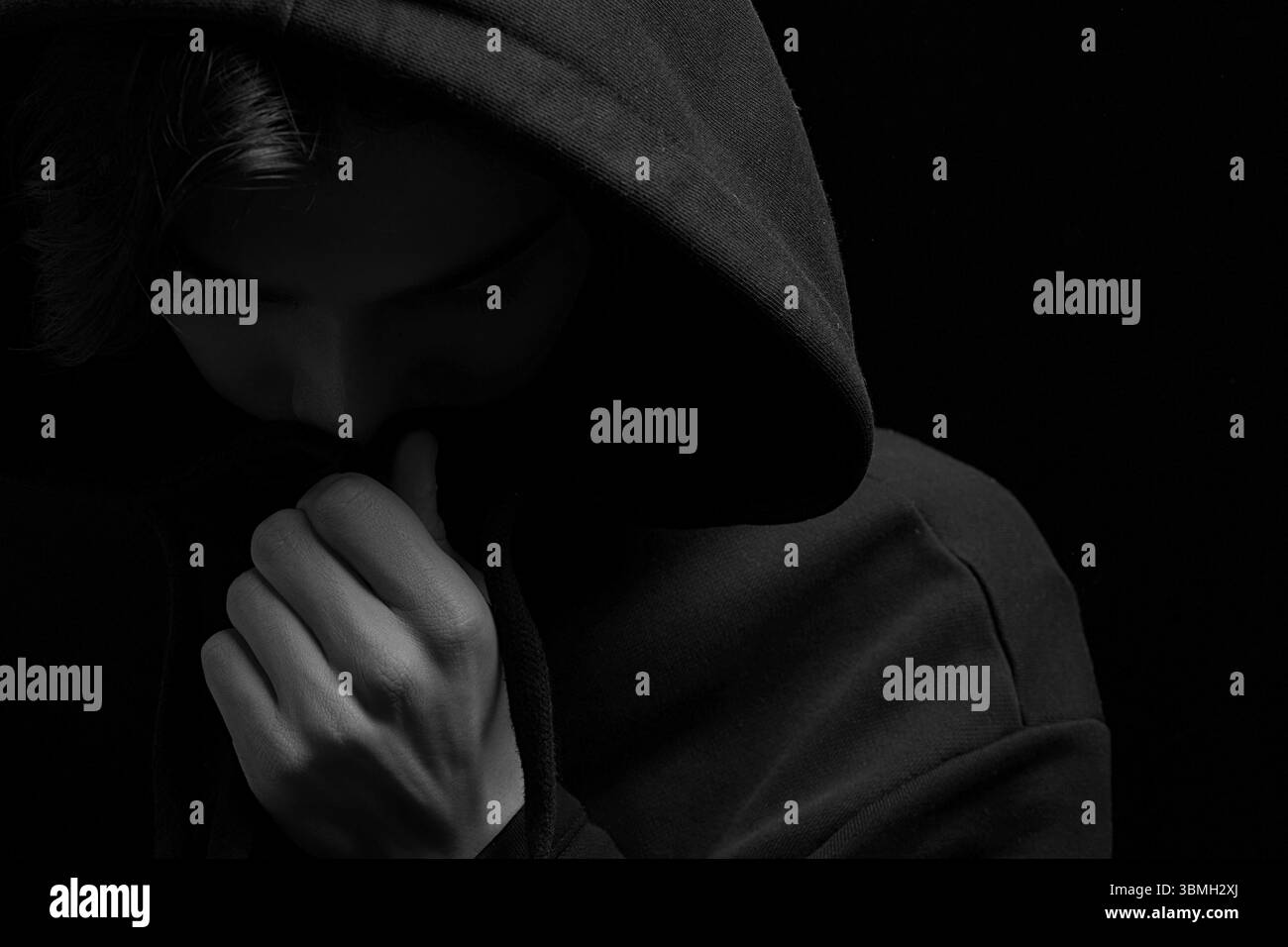 Ein junger kerl in einem schwarzen Hoodie auf einem schwarzen hintergrund mit seinem gesicht bedeckt Stockfoto