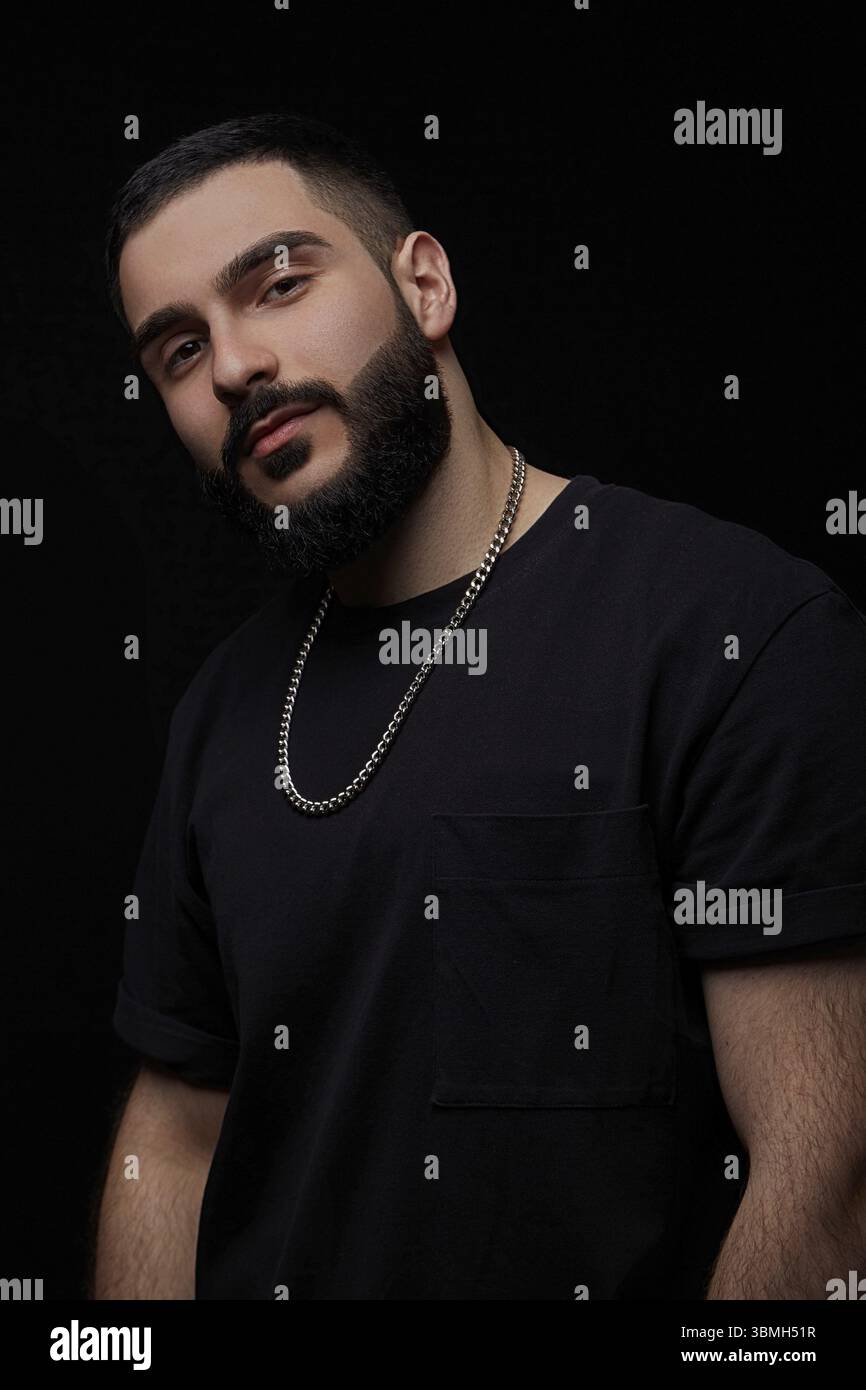 Ein dramatisches Porträt eines jungen seriösen Mannes, Musikers, Sängers, Rappers mit einem Bart und überkreuzten Armen auf der Brust auf einem schwarzen isolierten Hintergrundeer Stockfoto