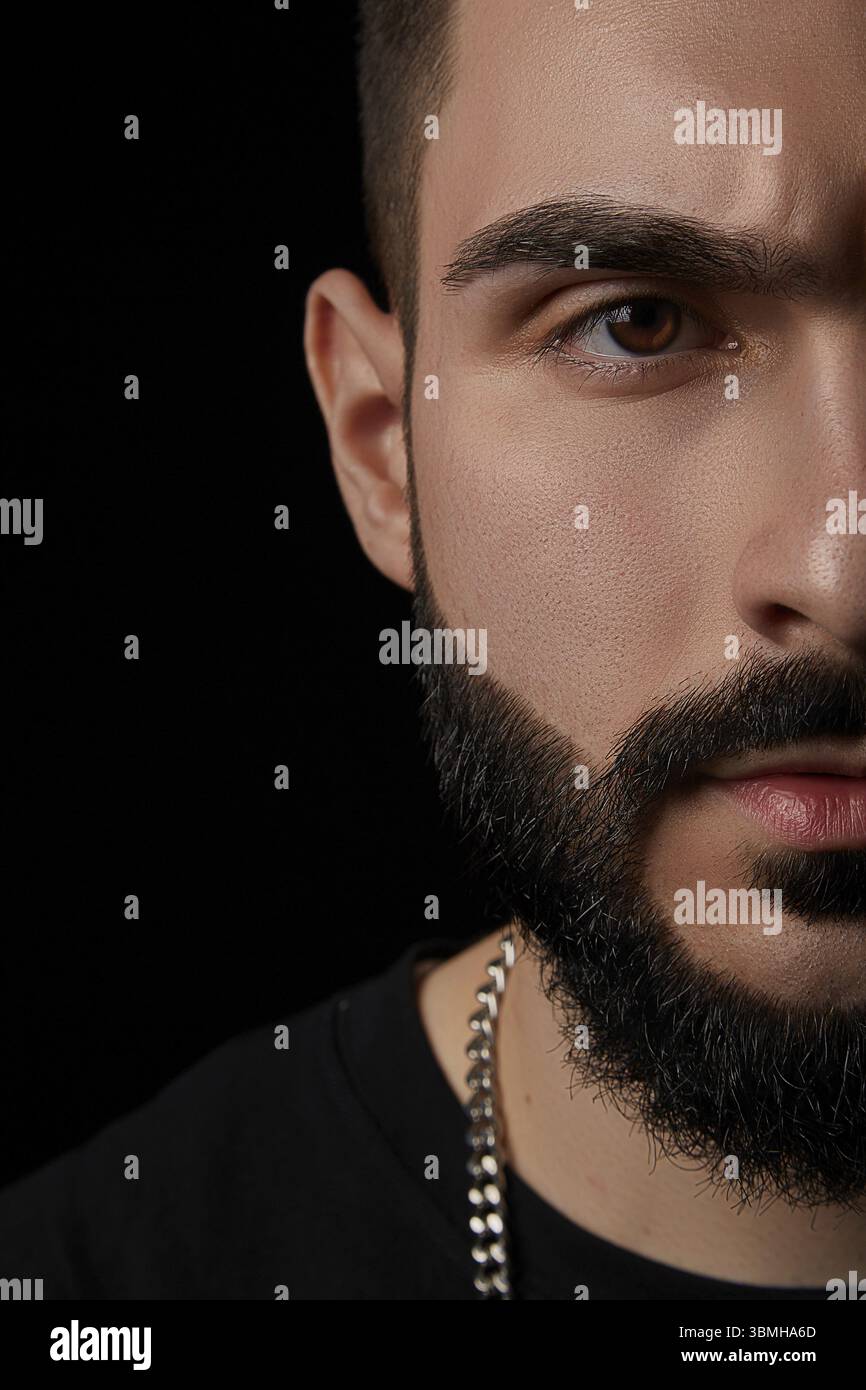Ein dramatisches Porträt eines jungen seriösen Mannes, Musikers, Sängers, Rappers mit einem Bart und überkreuzten Armen auf der Brust auf einem schwarzen isolierten Hintergrundeer Stockfoto