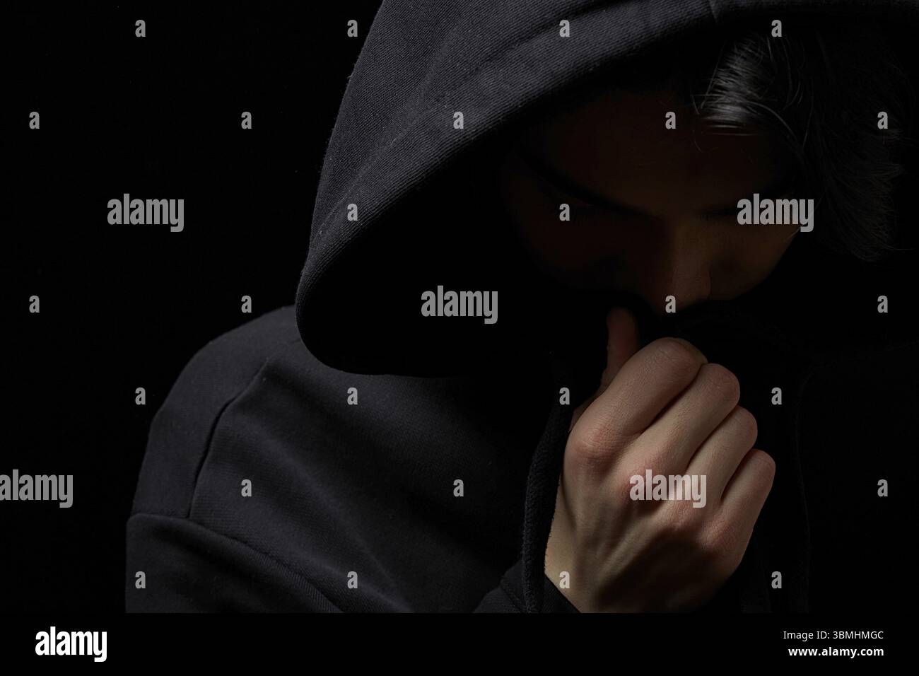 Ein junger kerl in einem schwarzen Hoodie auf einem schwarzen hintergrund mit seinem gesicht bedeckt Stockfoto