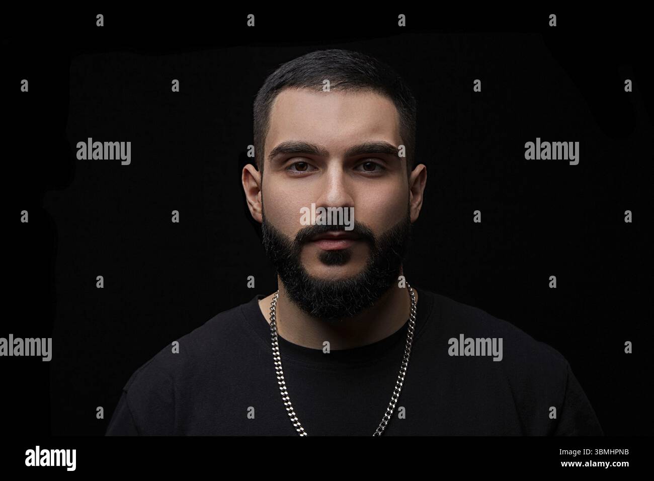 Ein dramatisches Porträt eines jungen seriösen Mannes, Musikers, Sängers, Rappers mit einem Bart und überkreuzten Armen auf der Brust auf einem schwarzen isolierten Hintergrundeer Stockfoto