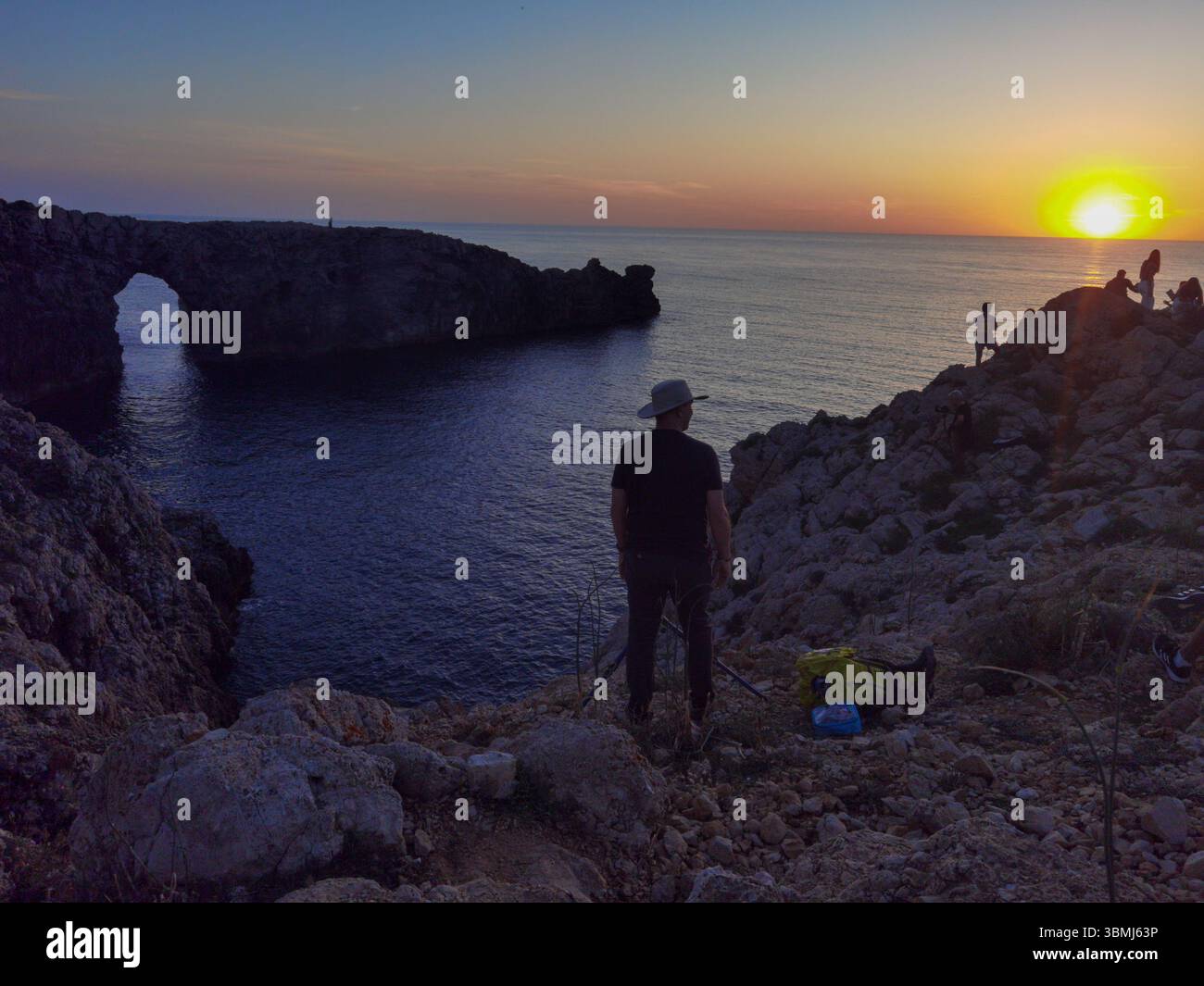 Fotograf, malerischer Sonnenuntergang, Meeresbogen. Ein Profi fängt die goldene Stunde über dem Pont d’en Gil auf Menorca ein, mit Silhouetten vor dramatischen Klippen und - Smartphone-aufgenommenes Stockfoto