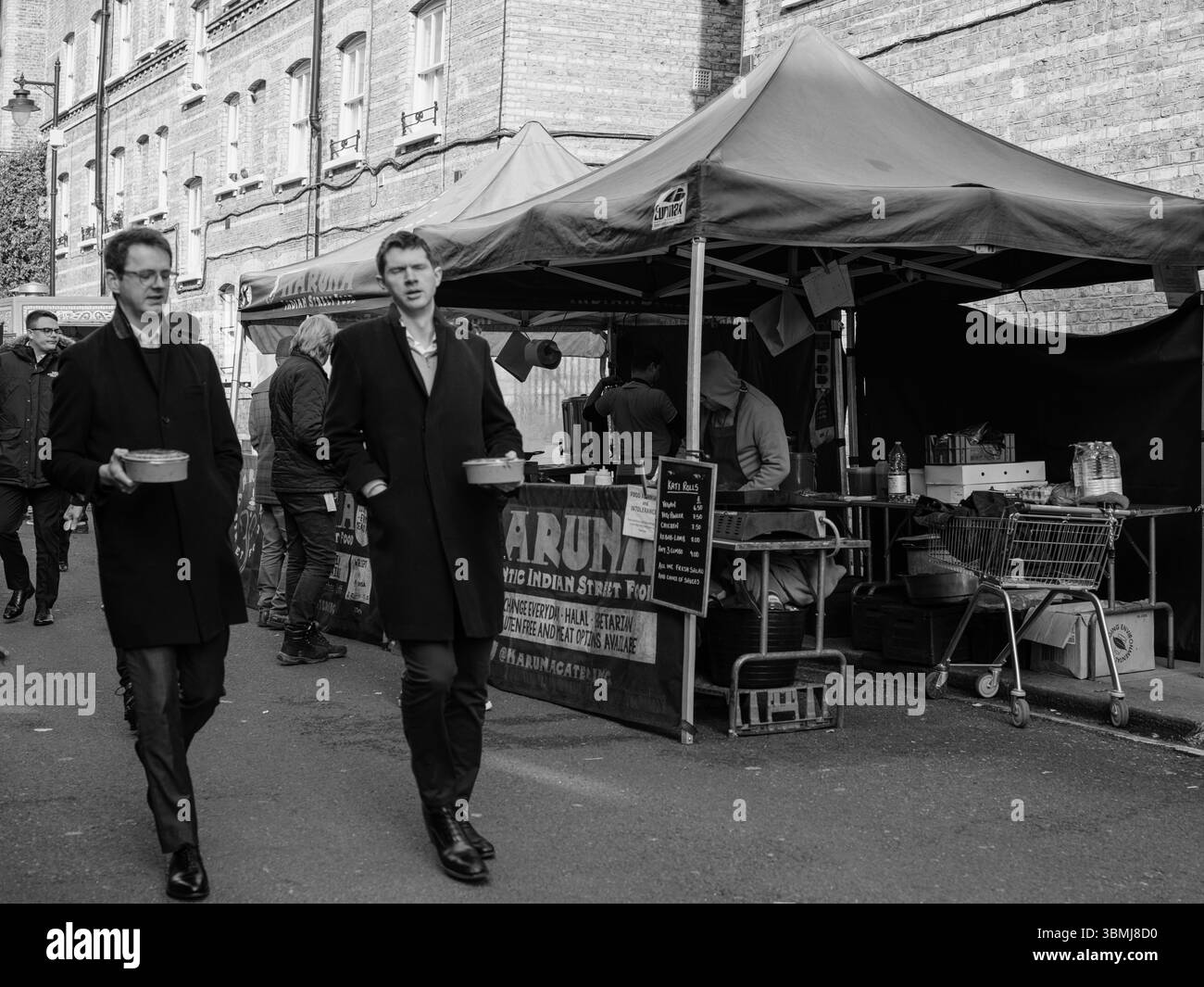 Vereinigtes Königreich, London, 4. Juli 2025. Schwarz-weiße Straßenszene, in der zwei Männer an einem Imbissstand auf einem Londoner Markt vorbeilaufen und täglich urbane Aufnahmen machen Stockfoto
