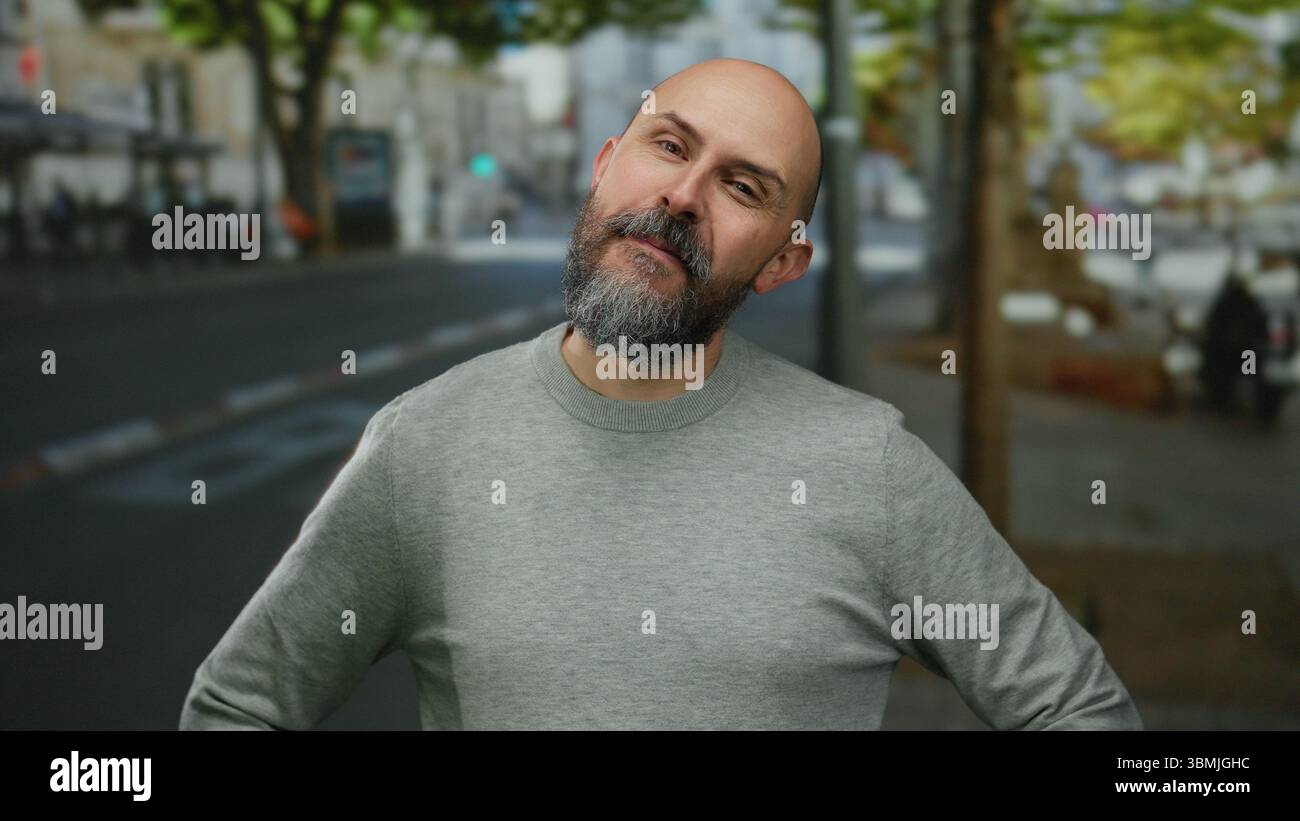 Bärtiger Mann in urbaner Umgebung mit grauem Pullover, der tagsüber auf einer von Bäumen gesäumten Stadtstraße warm lächelt und eine freundliche Outdoor-Atmosphäre darstellt. Stockfoto