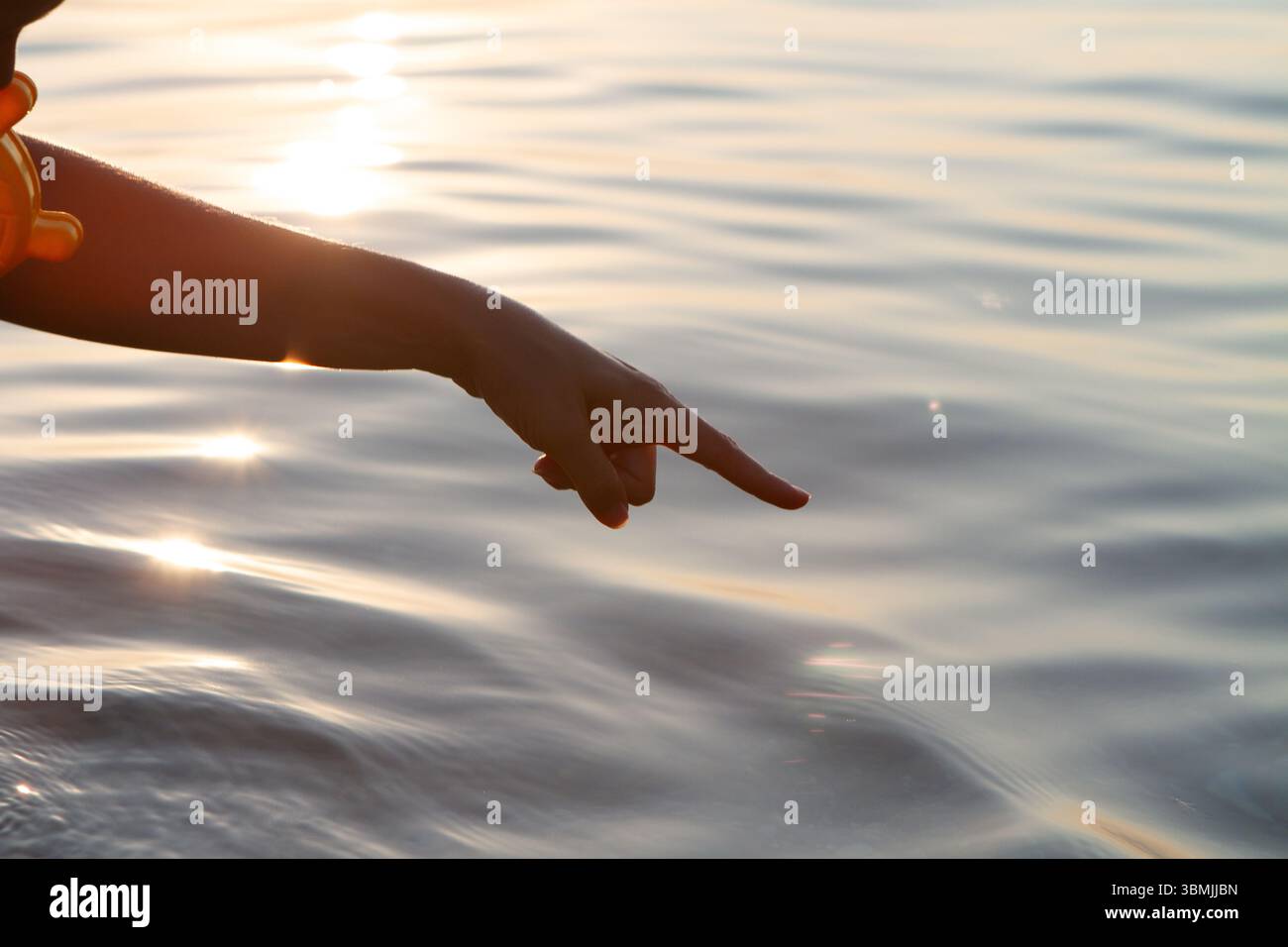 Hand zeigt bei Sonnenuntergang auf die Wasseroberfläche, mit Lichtreflexen, die eine warme und einladende Atmosphäre schaffen Stockfoto