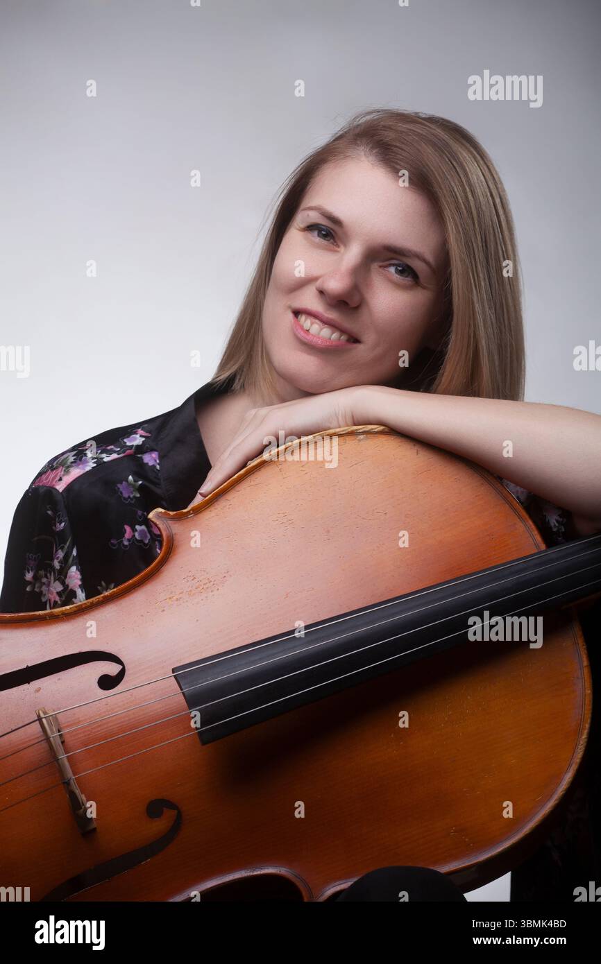 Junge kaukasische Frau, Ende 20/Anfang 30, lächelndes Atelierporträt mit Cello. Sie lehnt sich auf das Instrument und trägt ein schwarzes Blumenoberteil. Stockfoto