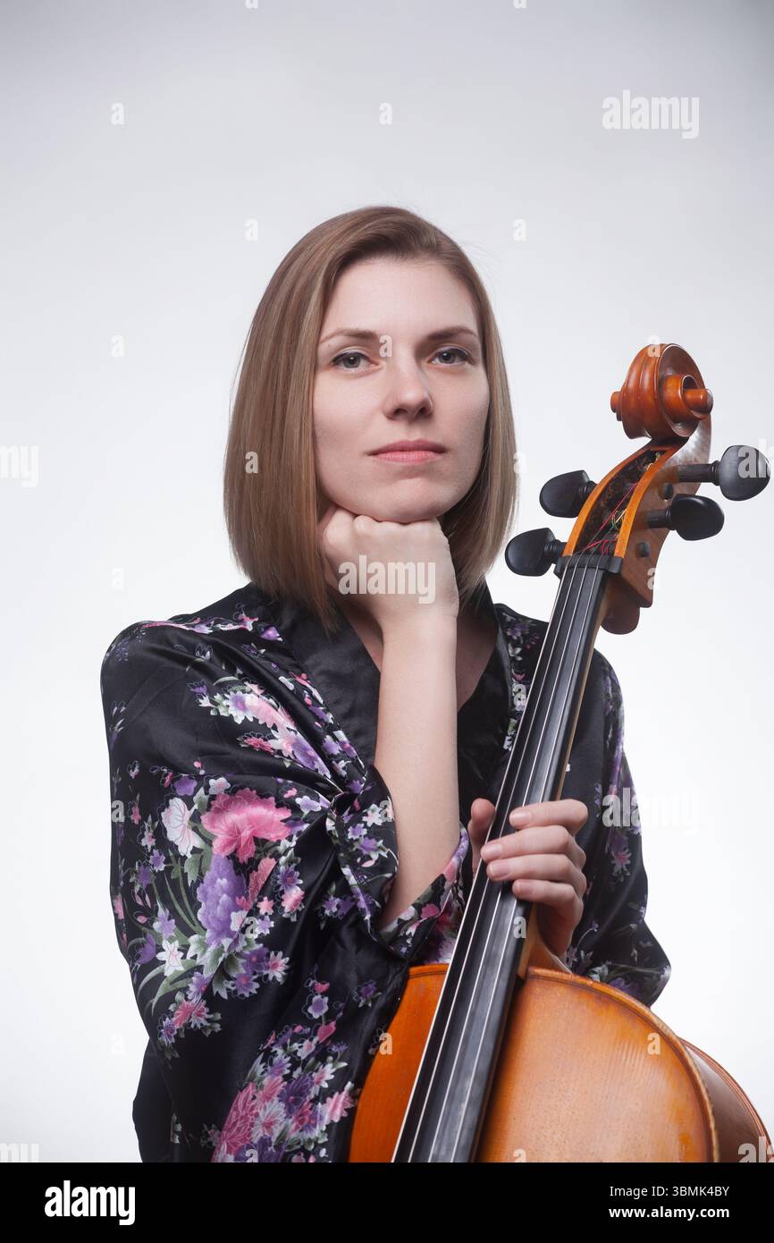 Erwachsene kaukasische Frau in schwarzem Blumengewand, die ein Cello in einem Atelierporträt hält, die Hand auf dem Kinn ruht. Stockfoto