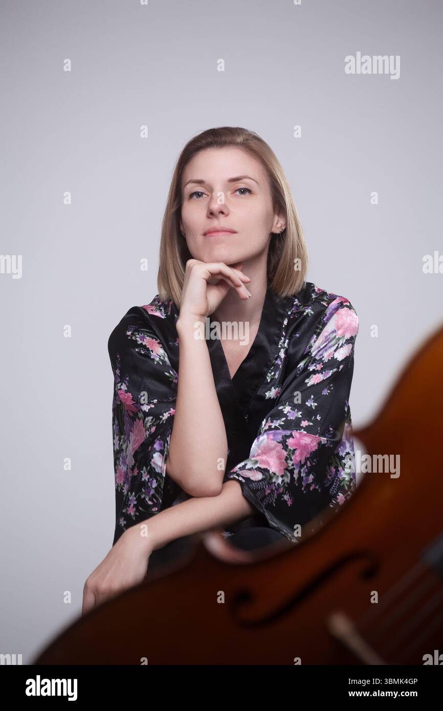 Kaukasische Frau, Ende 20 bis Anfang 30, in schwarzem Blumengewand, mit Hand am Kinn sitzend, nachdenklicher Ausdruck, Cello im Vordergrund, Studio-Aufnahme. Stockfoto