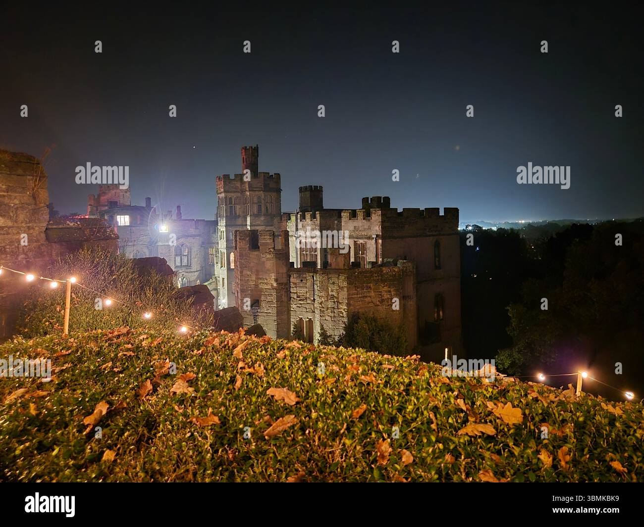 Beleuchtete historische Burggebäude bei Nacht mit dekorativen Lichtern und Herbstlaub im Vordergrund in Warwick, England. - Smartphone-aufgenommenes Stockfoto