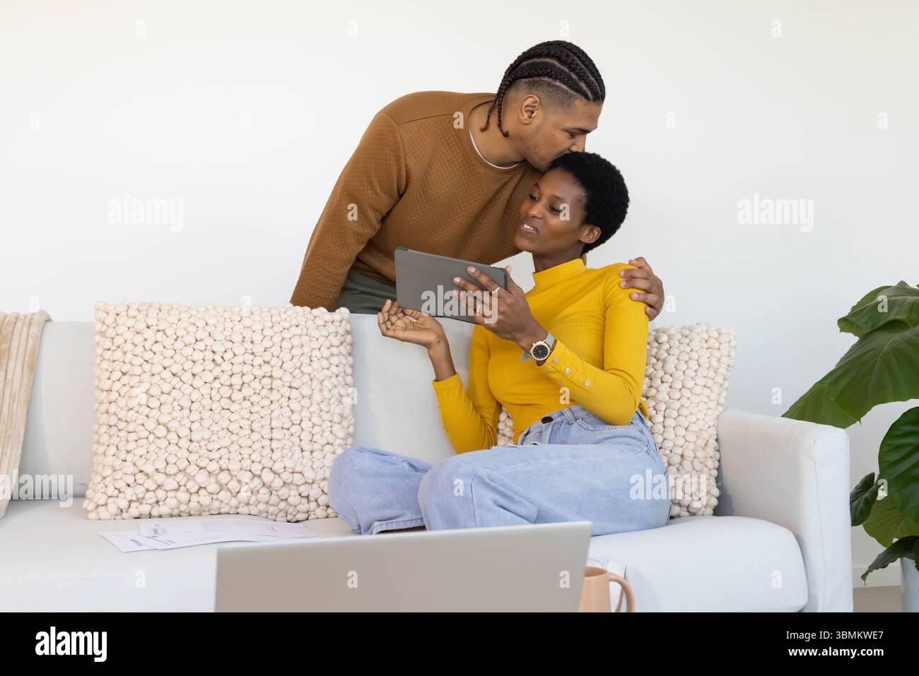 Küssendes Paar auf Sofa im Wohnzimmer, mit Tablet mit Laptop und Kaffeetasse Stockfoto