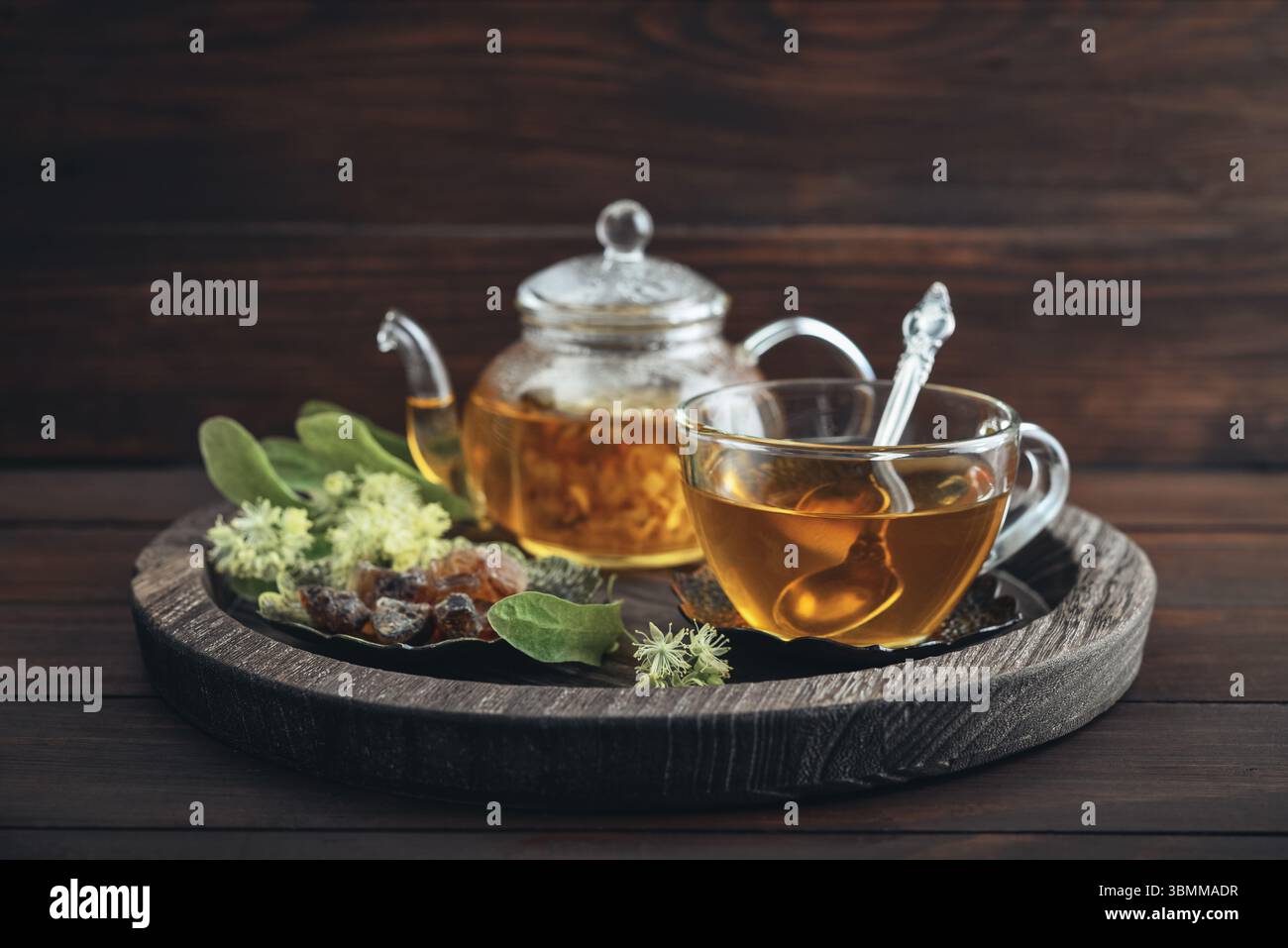 Glasschale Lindentee mit Glas-Teekanne, Zucker und Lindenblüten auf rundem Tablett auf hölzernem Hintergrund Stockfoto