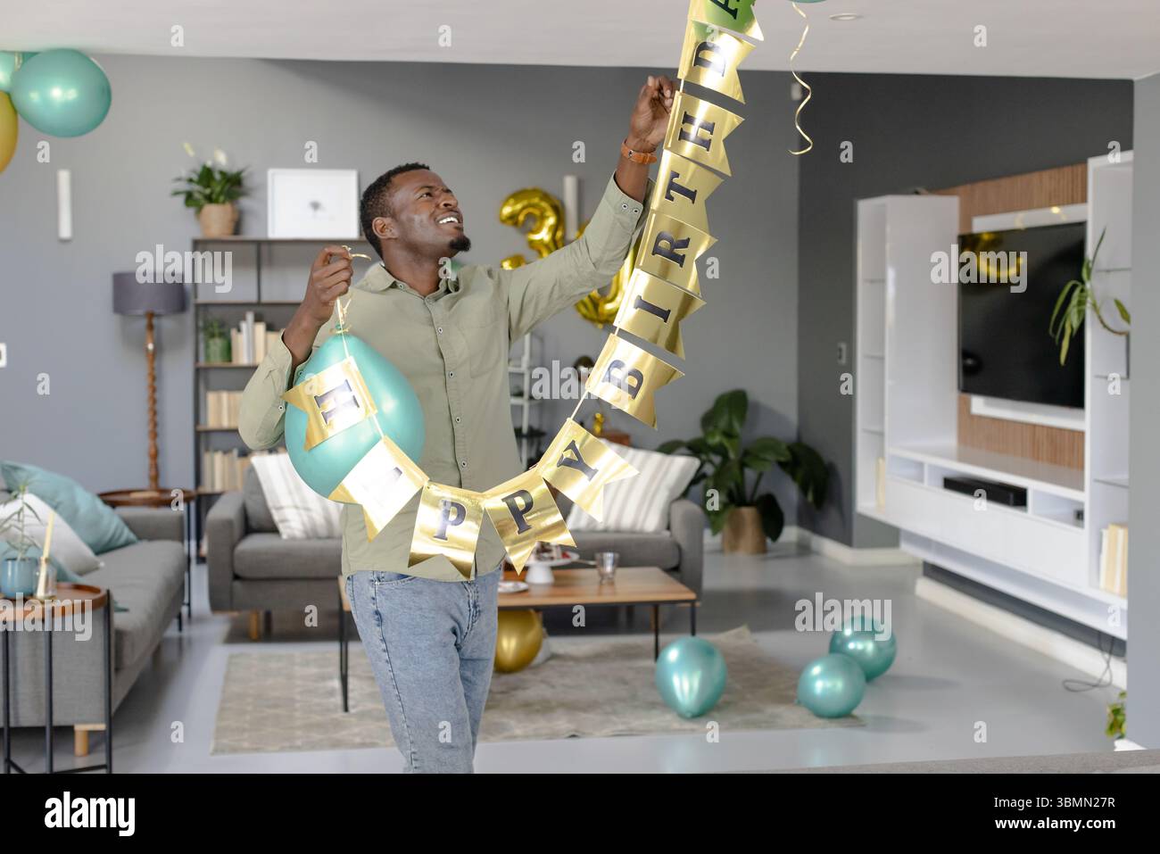 Afroamerikaner mit goldenem Happy Birthday Banner mit blauem Ballon im Wohnzimmer Stockfoto
