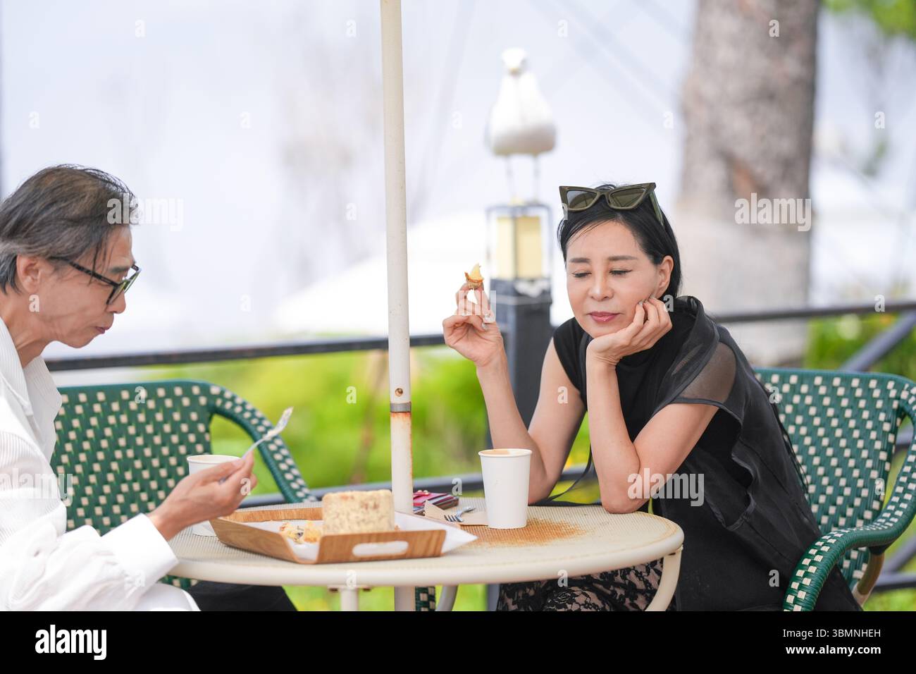 Auf der Terrasse eines Cafés am Meer genießen ein koreanischer Mann und eine koreanische Frau Ende der 50er Jahre Kaffee und Kuchen am Nachmittag, während eine Möwe aus der Nähe beobachtet und einen pe kreiert Stockfoto