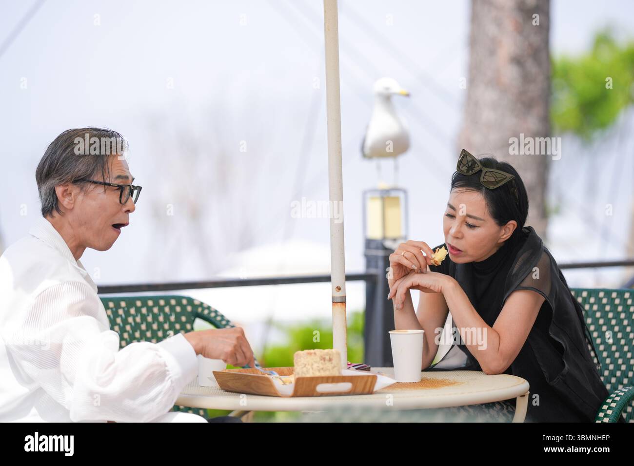 Auf der Terrasse eines Cafés am Meer genießen ein koreanischer Mann und eine koreanische Frau Ende der 50er Jahre Kaffee und Kuchen am Nachmittag, während eine Möwe aus der Nähe beobachtet und einen pe kreiert Stockfoto