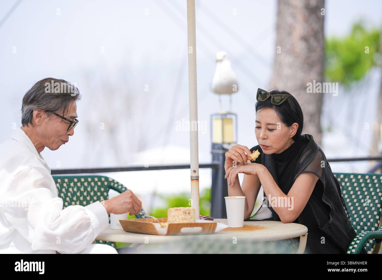 Auf der Terrasse eines Cafés am Meer genießen ein koreanischer Mann und eine koreanische Frau Ende der 50er Jahre Kaffee und Kuchen am Nachmittag, während eine Möwe aus der Nähe beobachtet und einen pe kreiert Stockfoto