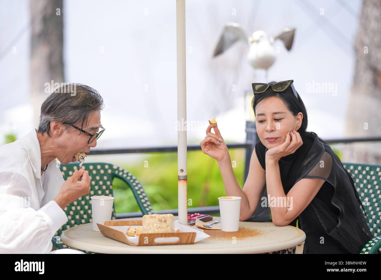 Auf der Terrasse eines Cafés am Meer genießen ein koreanischer Mann und eine koreanische Frau Ende der 50er Jahre Kaffee und Kuchen am Nachmittag, während eine Möwe aus der Nähe beobachtet und einen pe kreiert Stockfoto