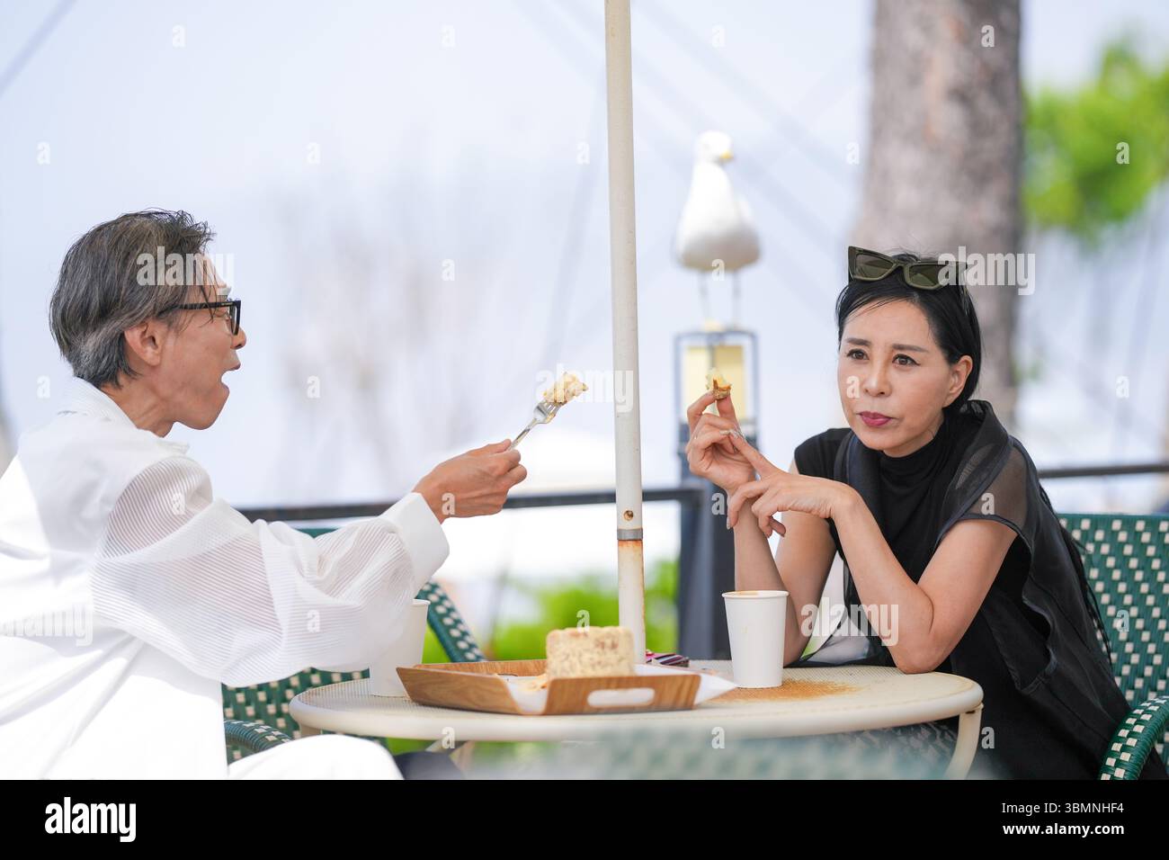Auf der Terrasse eines Cafés am Meer genießen ein koreanischer Mann und eine koreanische Frau Ende der 50er Jahre Kaffee und Kuchen am Nachmittag, während eine Möwe aus der Nähe beobachtet und einen pe kreiert Stockfoto