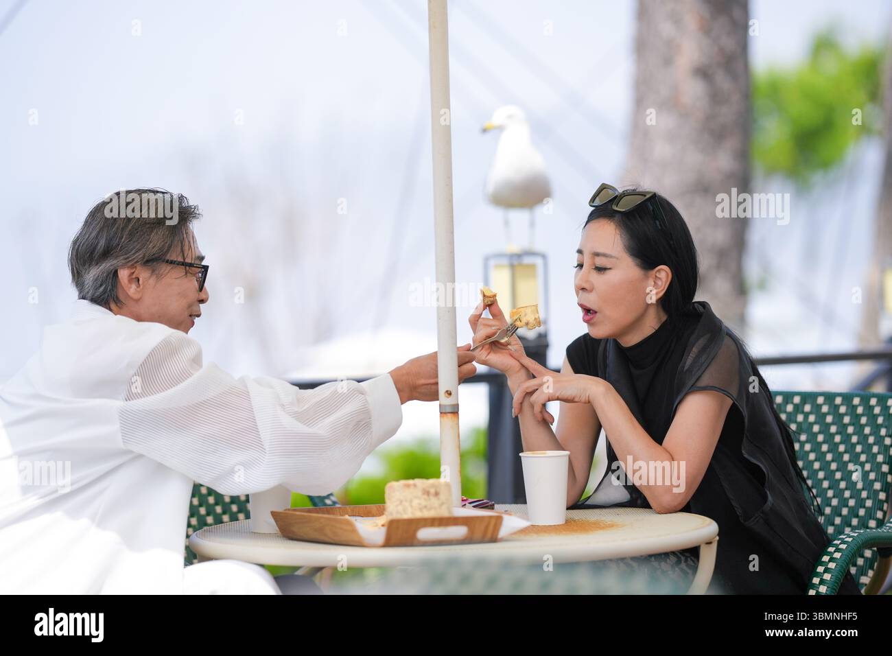 Auf der Terrasse eines Cafés am Meer genießen ein koreanischer Mann und eine koreanische Frau Ende der 50er Jahre Kaffee und Kuchen am Nachmittag, während eine Möwe aus der Nähe beobachtet und einen pe kreiert Stockfoto