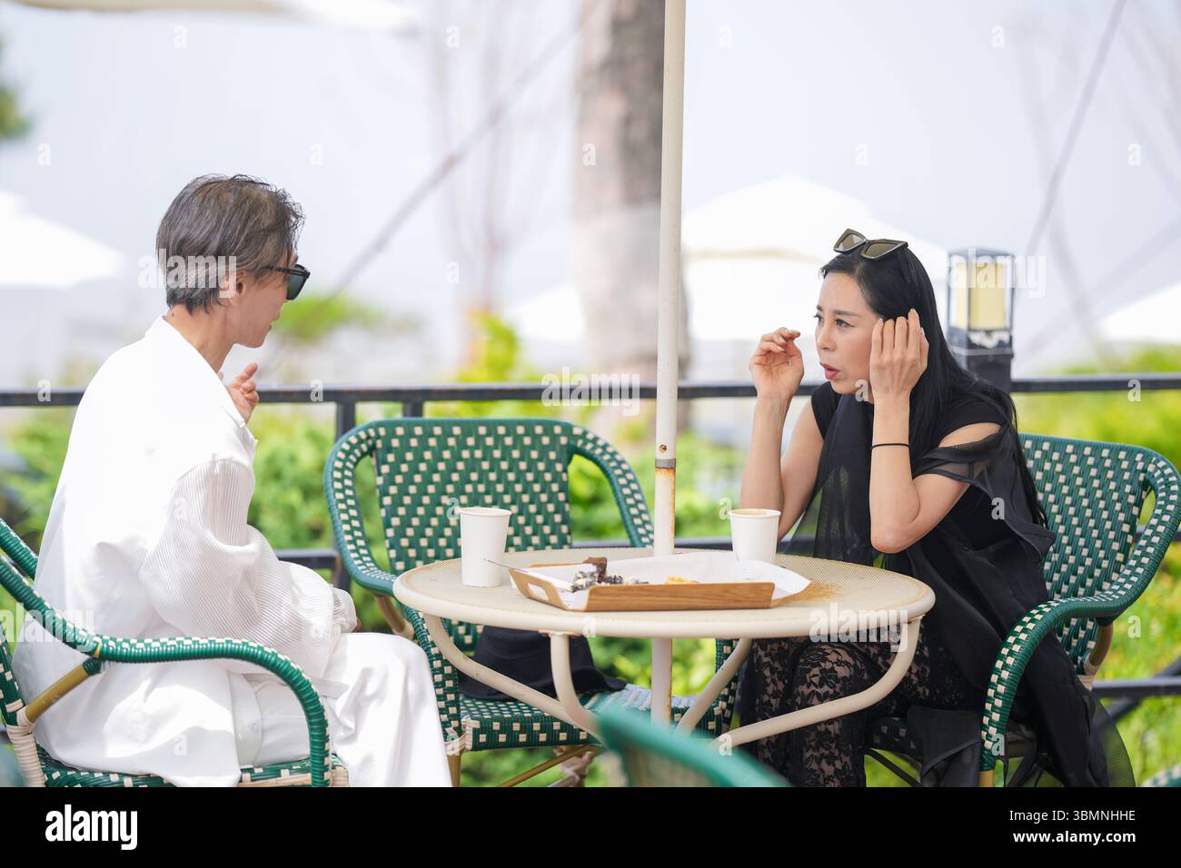 Auf der Terrasse eines Cafés am Meer genießen ein koreanischer Mann und eine koreanische Frau Ende der 50er Jahre Kaffee und Kuchen am Nachmittag, während eine Möwe aus der Nähe beobachtet und einen pe kreiert Stockfoto