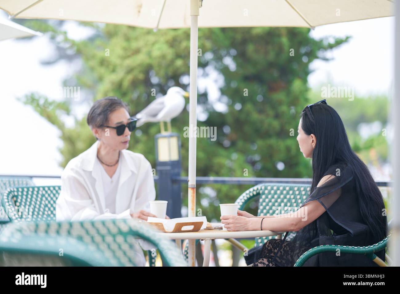 Auf der Terrasse eines Cafés am Meer genießen ein koreanischer Mann und eine koreanische Frau Ende der 50er Jahre Kaffee und Kuchen am Nachmittag, während eine Möwe aus der Nähe beobachtet und einen pe kreiert Stockfoto