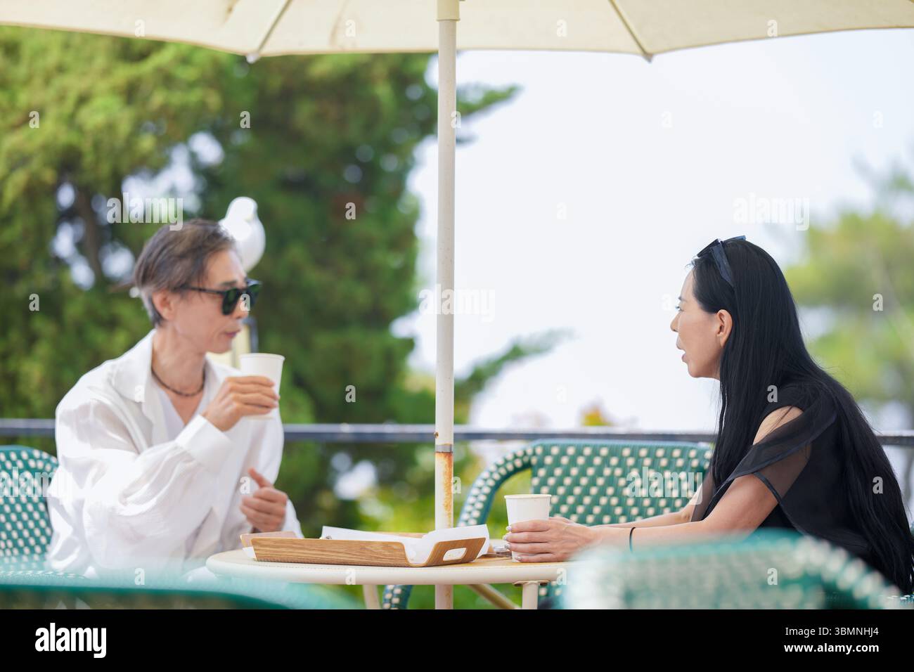 Auf der Terrasse eines Cafés am Meer genießen ein koreanischer Mann und eine koreanische Frau Ende der 50er Jahre Kaffee und Kuchen am Nachmittag, während eine Möwe aus der Nähe beobachtet und einen pe kreiert Stockfoto
