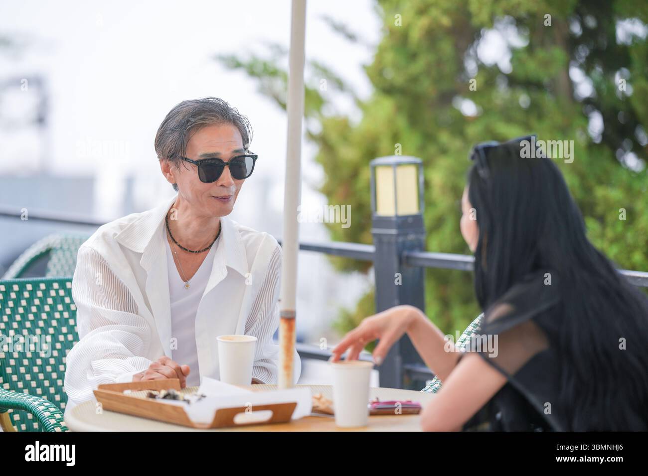 Auf der Terrasse eines Cafés am Meer genießen ein koreanischer Mann und eine koreanische Frau Ende der 50er Jahre Kaffee und Kuchen am Nachmittag, während eine Möwe aus der Nähe beobachtet und einen pe kreiert Stockfoto