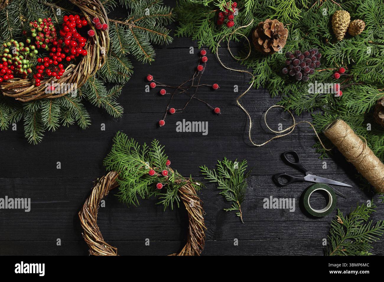 Machen Sie einen Weihnachtskranz mit Ihren eigenen Händen. Fichtenzweig, Weihnachtskranz und Geschenke auf schwarzem Holzhintergrund. Arbeitsplatz für die Zubereitung von handgefertigten Stockfoto