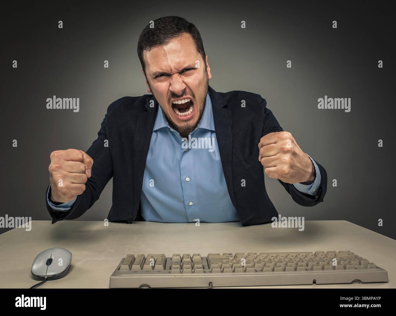 Porträt eines wütenden, verärgerten jungen Mannes in blauem Hemd und Jacke mit erhobenen Fäusten, der an einem Schreibtisch in der Nähe eines Computers sitzt, isoliert auf grauem Hintergrund. Negati Stockfoto