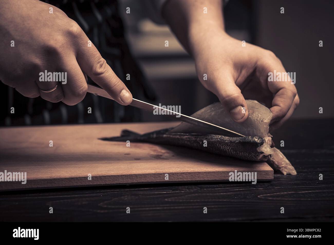Koch schneidet den Fisch auf einem Brett. Foto in Brauntönen. Küche. Holztisch. Hände des Kochs aus der Nähe Stockfoto