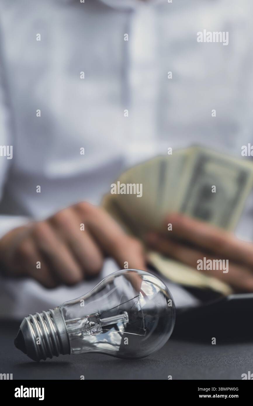 Elektrische Glühlampe. Der Mann übergibt Zählkosten für Elektrizitätsscheine und Bargeldbuchungen in Dollar an den Rechner. Nahaufnahme der Hände, die nicht erkennbar sind Stockfoto