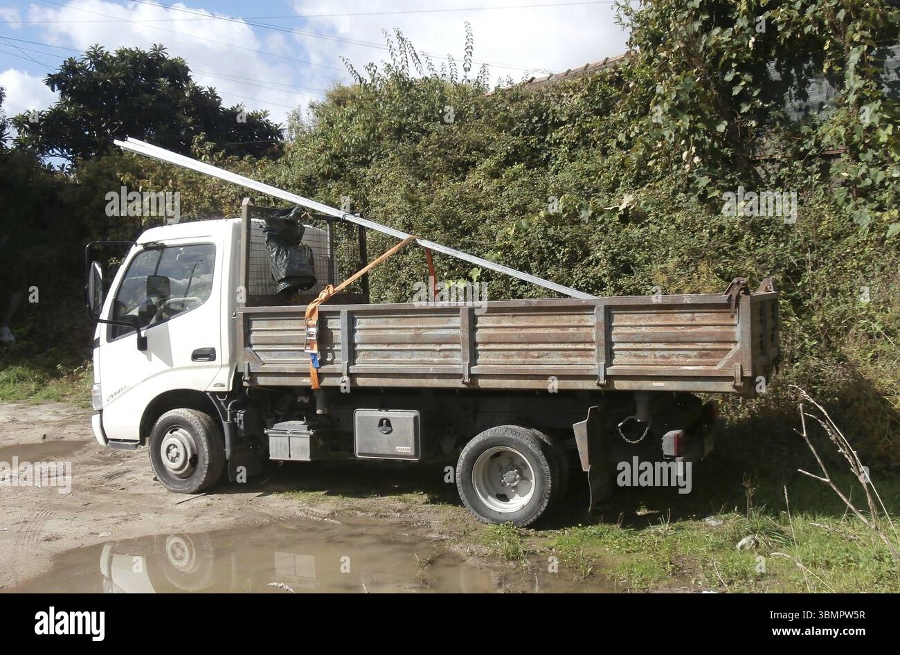 Weißer Toyota Dyna Truck, der lange Metallträger transportiert, die mit Gurten auf einer unbefestigten Straße gesichert sind Stockfoto