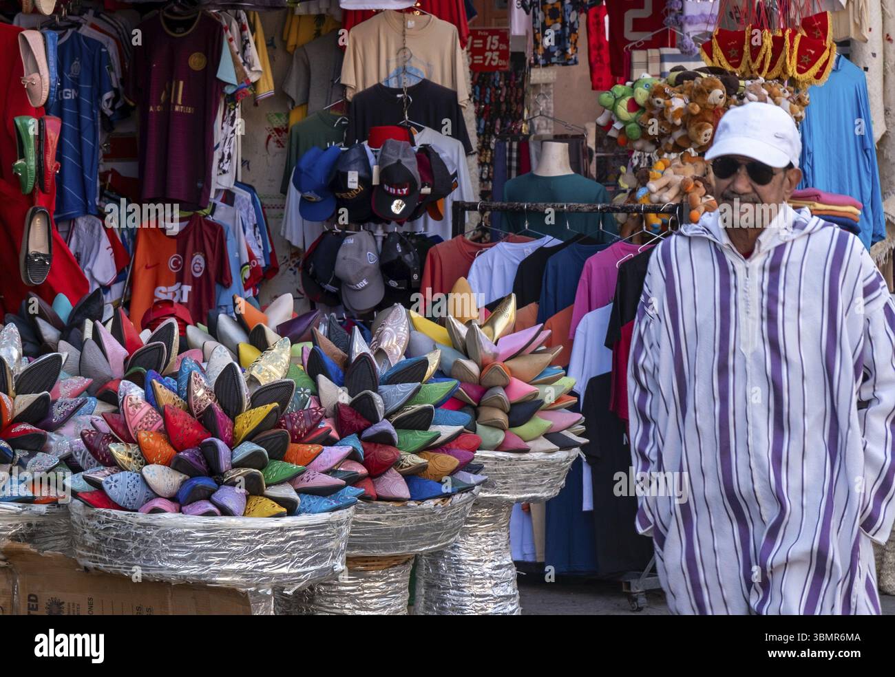 Straßenverkäufer mit Djellaba, der neben bunten Babouche-Hausschuhen und Kleidung auf einem traditionellen marokkanischen Markt steht Stockfoto