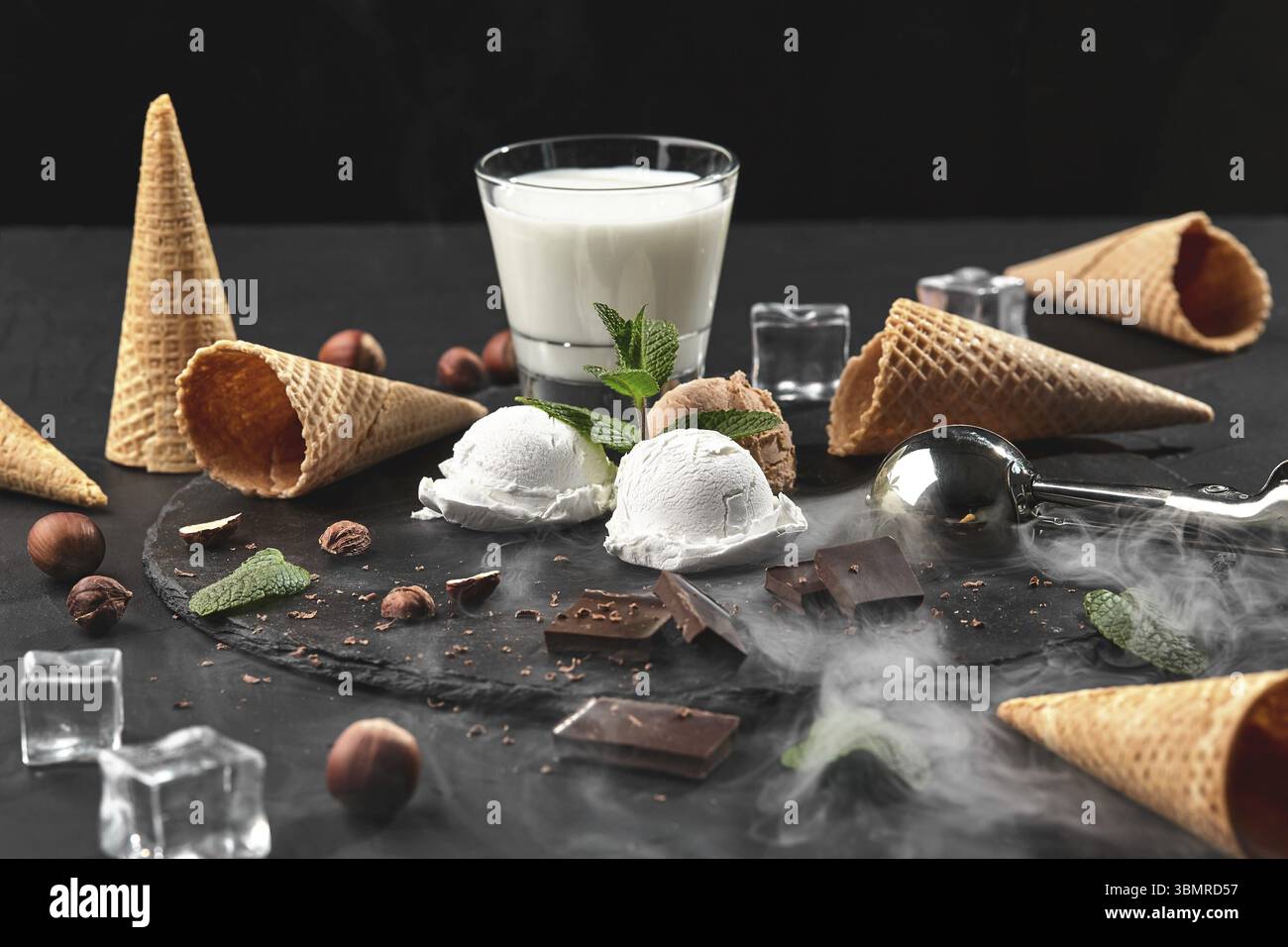 Es werden Schokoladen- und cremige Eiskugeln serviert, die mit Minze dekoriert sind, ein Glas Milch, Eiswürfel, Waffelkegel mit Schokolade und Haselnüssen in der Nähe Stockfoto