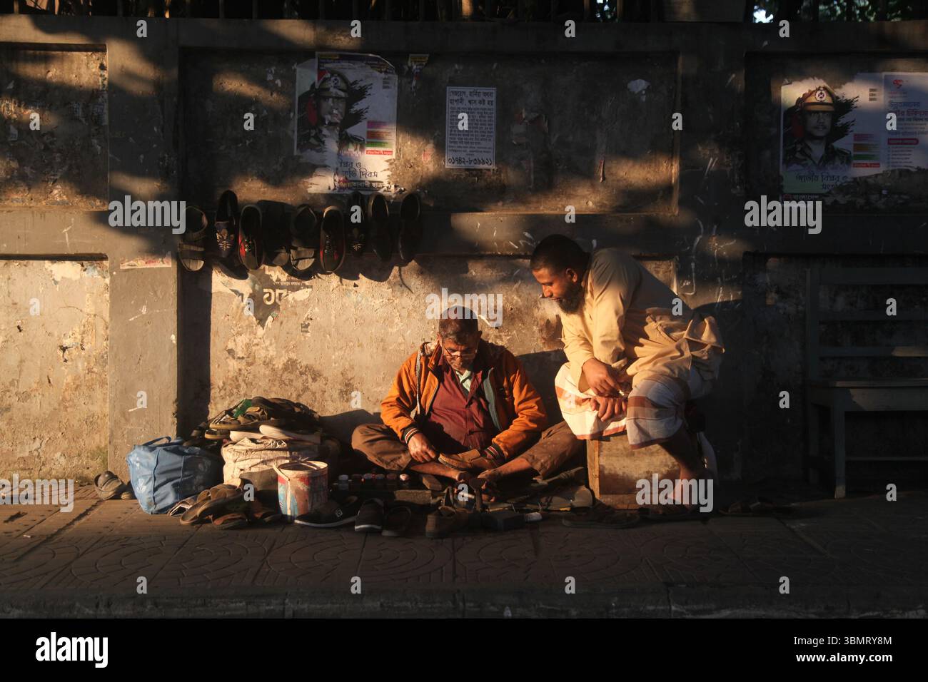 Dhaka Bangladesch 27. Dezember 2024, der Kampf des Lebens zwischen Licht und Schatten , Ein Schuster repariert Schuhe auf dem Bürgersteig, während ein Kunde sitzt Stockfoto