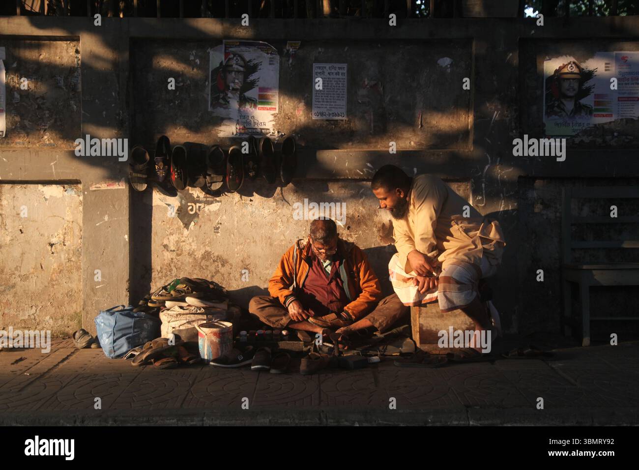 Dhaka Bangladesch 27. Dezember 2024, der Kampf des Lebens zwischen Licht und Schatten , Ein Schuster repariert Schuhe auf dem Bürgersteig, während ein Kunde sitzt Stockfoto