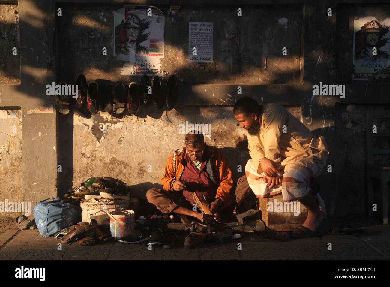 Dhaka Bangladesch 27. Dezember 2024, der Kampf des Lebens zwischen Licht und Schatten , Ein Schuster repariert Schuhe auf dem Bürgersteig, während ein Kunde sitzt Stockfoto