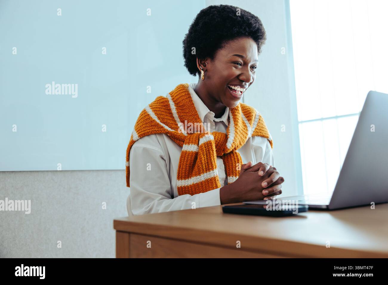 Eine fröhliche Geschäftsfrau wird während eines virtuellen Meetings mit einem warmen Lächeln dargestellt. Sie trägt einen leuchtenden orangefarbenen Pullover, der über einem weißen Hemd hängt Stockfoto