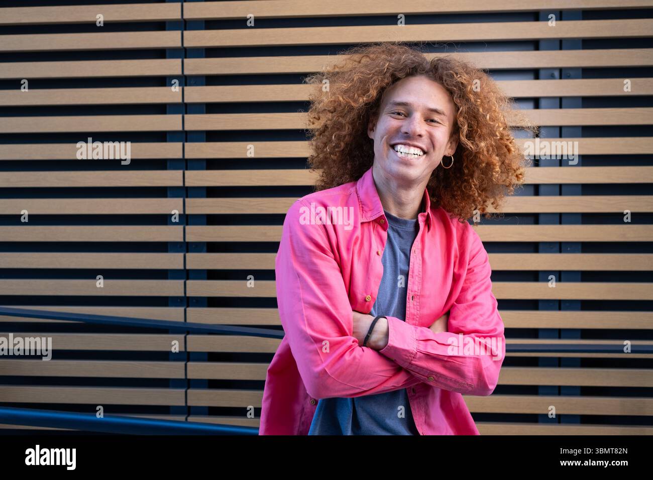 Porträt eines fröhlichen jungen Mannes mit lockigen Haaren, der selbstbewusst lächelt und die Arme vor der modernen Holzwand gekreuzt hat. Lebendiger Look, lässige Mode, urb Stockfoto