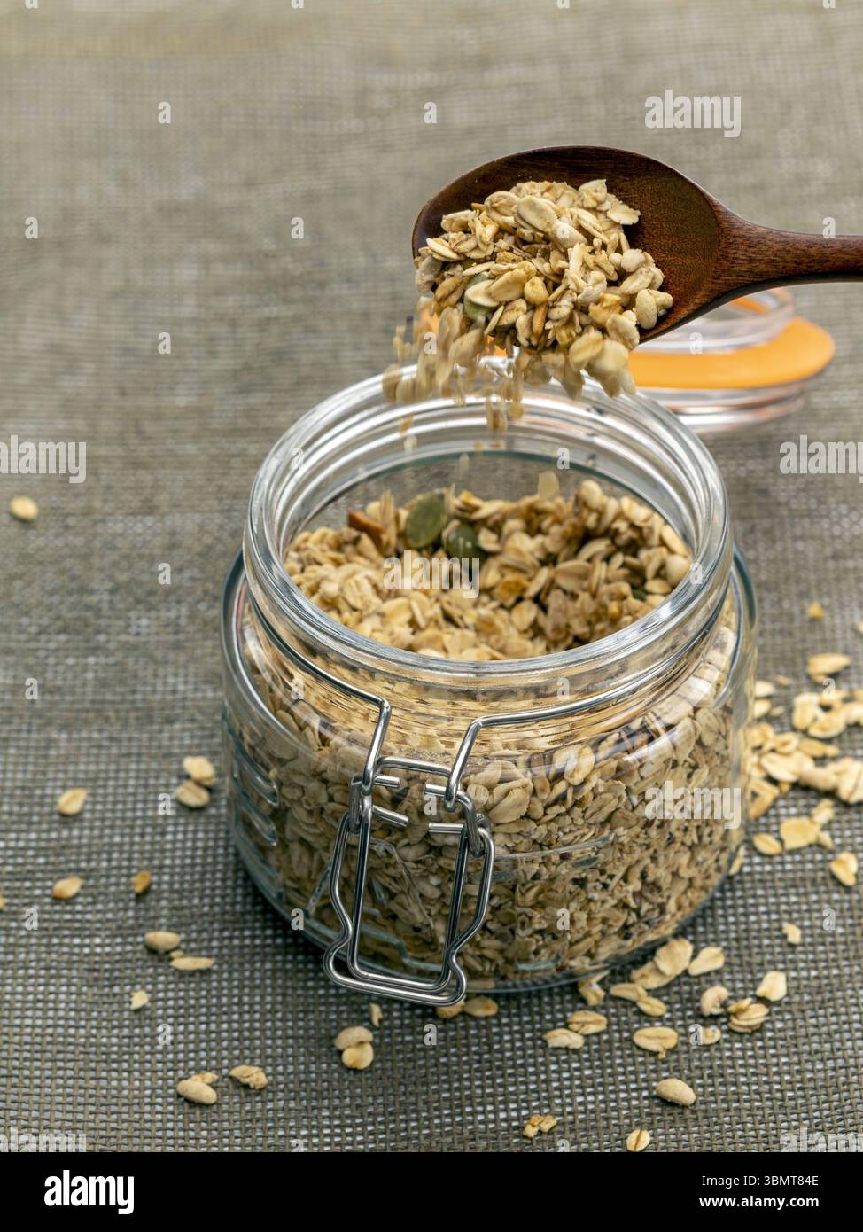 Ein mit trockenem Haferflocken gefülltes Gefäß wird mit einem Holzlöffel geschaufelt, wodurch ein Teil Hafer auf eine strukturierte Oberfläche verschüttet wird. Szene vermittelt rustikales und gesundes Leben Stockfoto