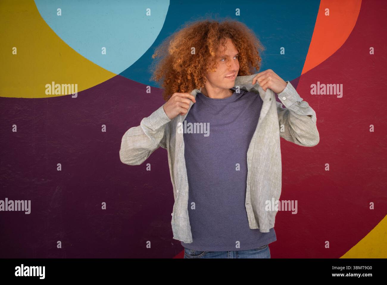 Junger Mann mit lockigem Haar, der sein Hemd vor einer bunten abstrakten Wand anpasst. Urbane Mode, lässiger Stil, lebendiger Hintergrund, Jugendstil, Stockfoto