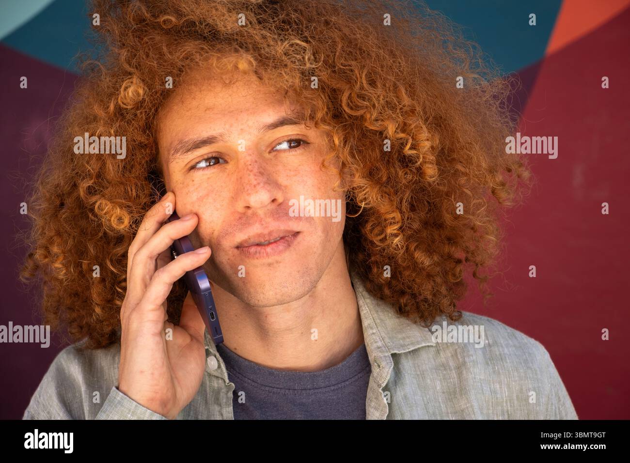 Nahaufnahme eines jungen Schülers mit lockigen Haaren, der auf dem Smartphone spricht, nachdenklicher Ausdruck vor einem farbenfrohen Wandgemälde. Urbane Umgebung, Universitätsleben Stockfoto