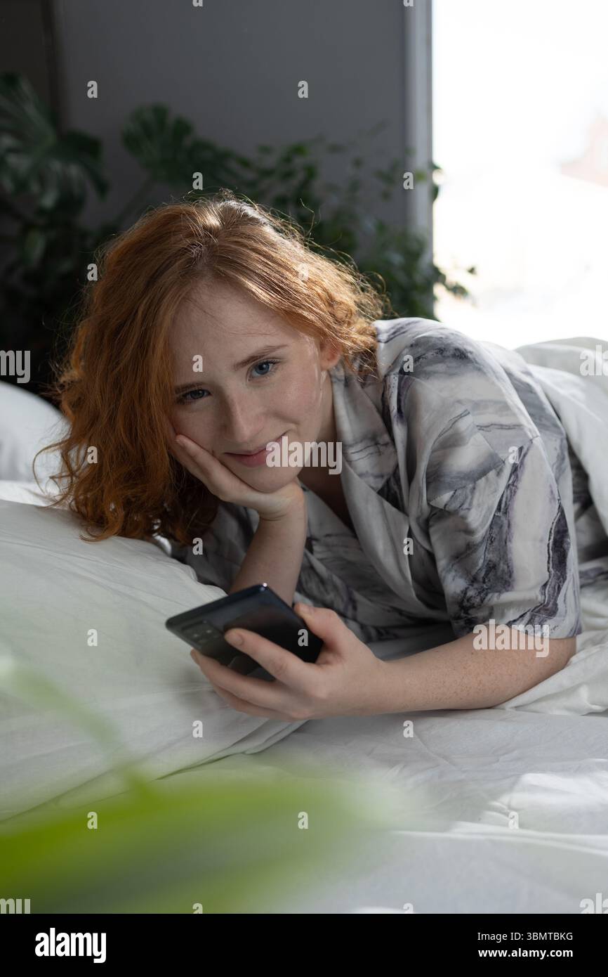 Eine junge Frau genießt einen glücklichen Moment im Bett, lächelt mit ihrem Smartphone, umgeben von Grün und Licht Stockfoto