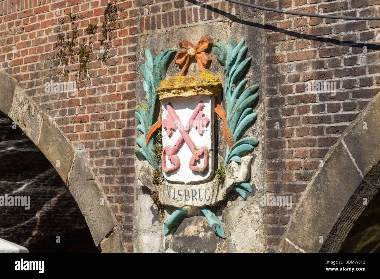 Ein historisches Wappen mit gekreuzten Schlüsseln und einem Band, das vor einer verwitterten Backsteinbrücke steht und Leiden' reiche Vergangenheit und die „Visbrug“ symbolisiert. Stockfoto