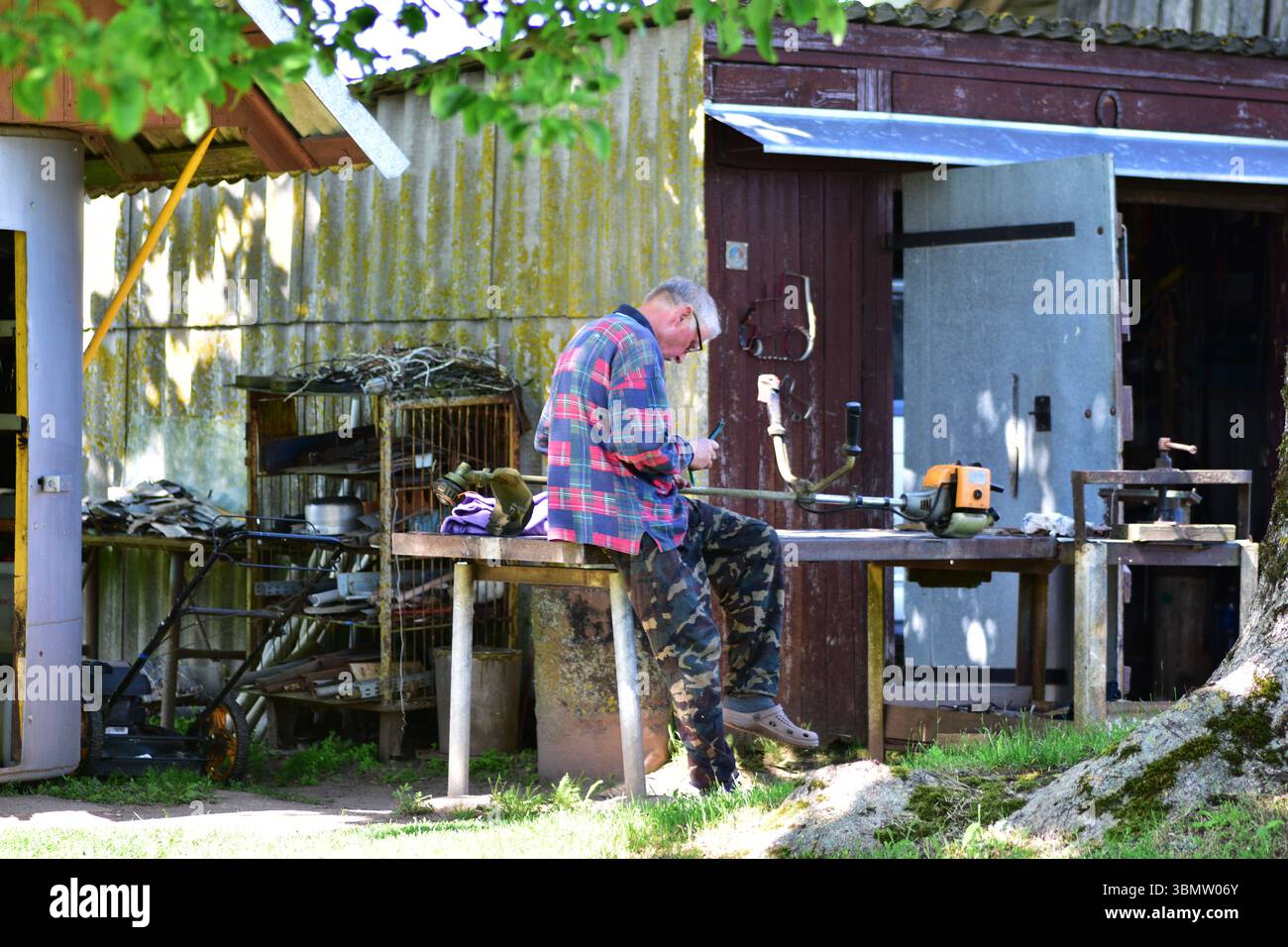Älterer Mann in kariertem Hemd und Camo-Hose, der an Gartengeräten vor dem rustikalen Schuppen arbeitet. Sommer-Landschaft mit Werkstattwerkzeugen und Natur-SHA Stockfoto