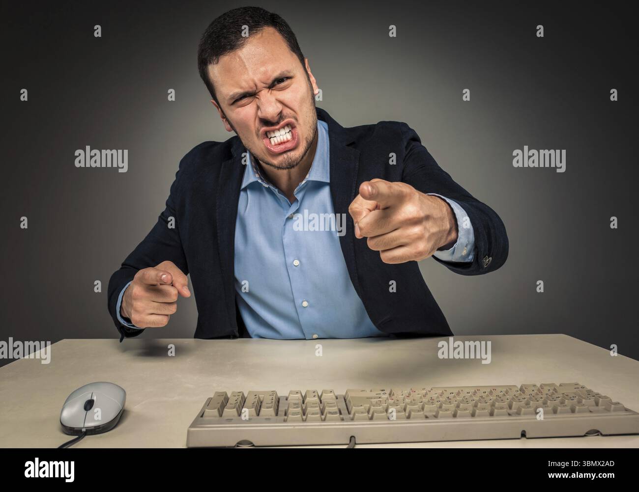 Porträt eines wütenden, verärgerten jungen Mannes in blauem Hemd und Jacke mit erhobenen Händen, der an einem Schreibtisch in der Nähe eines Computers sitzt, isoliert auf grauem Hintergrund. Negati Stockfoto